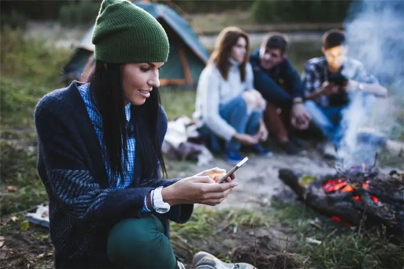 Jauna moteris naudojasi mobiliuoju telefonu, kai draugai sėdi prie laužo stovyklavietėje